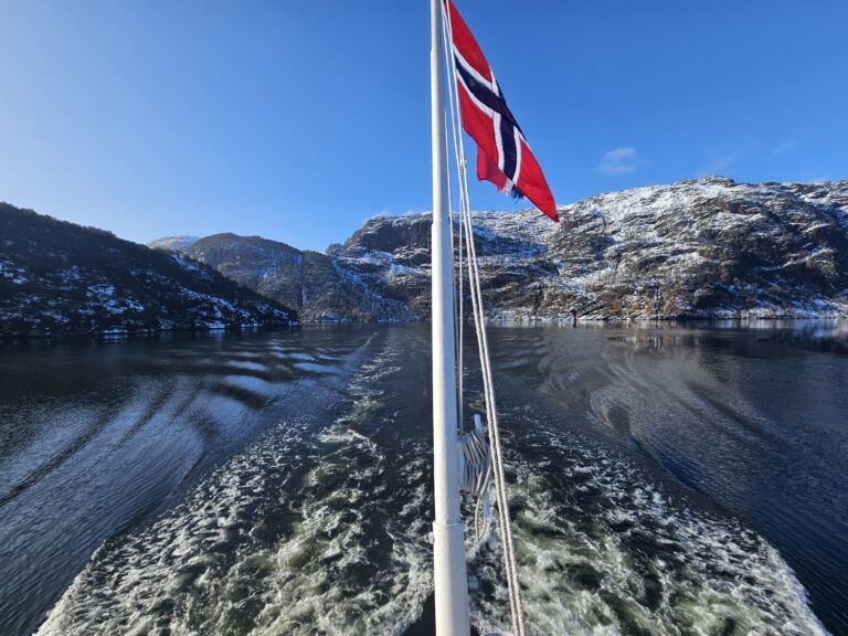 Back of the Mostraumen Fjord Cruise on a sunny day, with Norwegian flag and fjords in the background.